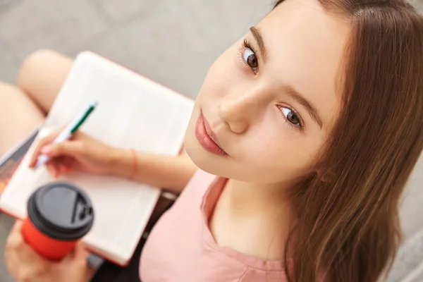 Escola menina escrevendo caneta no caderno sobre os joelhos. Estudante com café em uma xícara de papel perto da universidade . — Fotografia de Stock