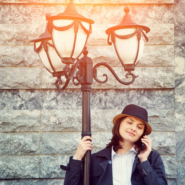 一个戴着帽子，戴着太阳镜，手持智能手机的时髦女人站在灯柱前的户外肖像. — 图库照片