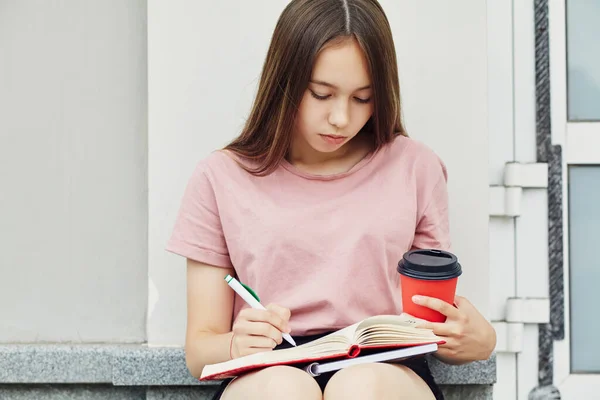 Дівчина пише ручку в блокноті. Студент з кавою в паперовій чашці біля університету . — стокове фото