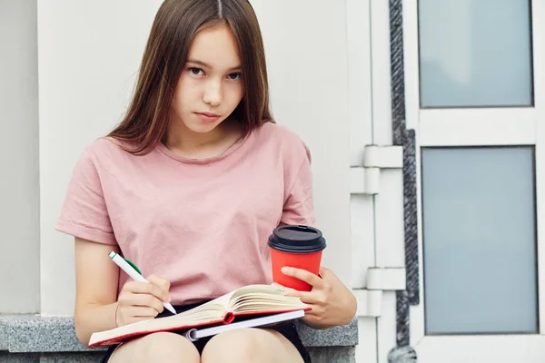 Holka píše pero do sešitu. Student s kávou v papírovém kelímku u univerzity. — Stock fotografie