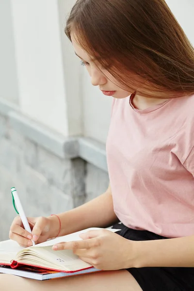 Школярка пише ручку в блокноті на колінах. Студент біля університету . — стокове фото