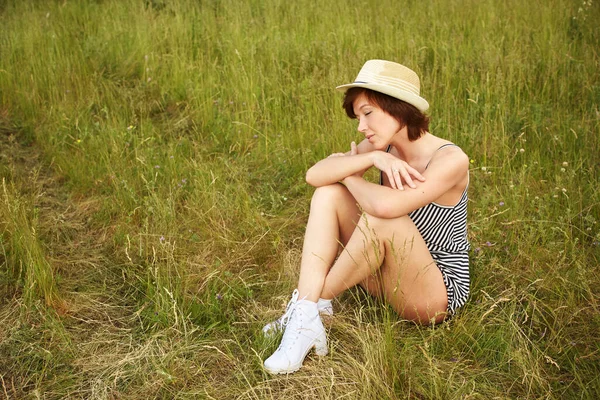 Retrato de verão de uma jovem mulher em um chapéu sentado em uma grama de prado . — Fotografia de Stock