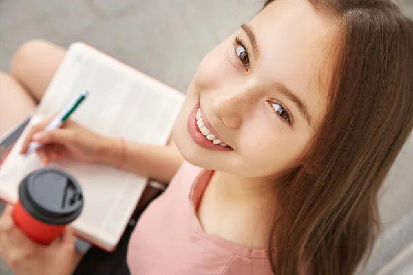 Девушка пишет ручку в блокноте. Студент с кофе в бумажной чашке возле университета — стоковое фото