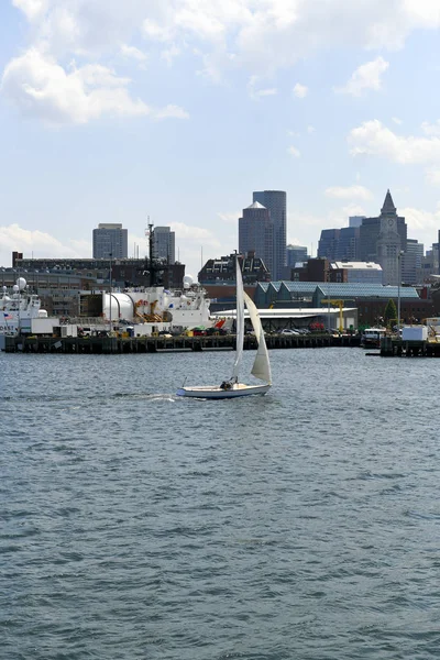 A small sailboat passes by a large docked Coast Guard vessel along the Boston Harbor shoreline.