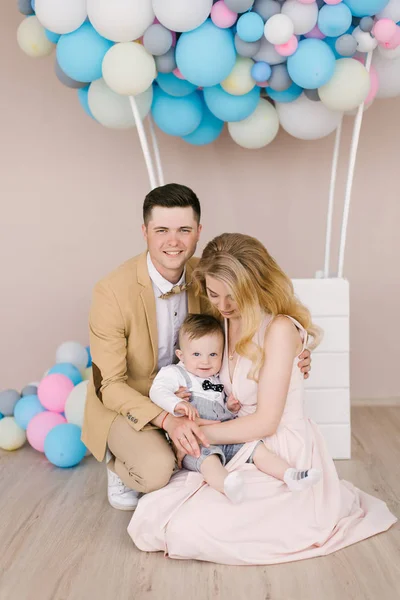 Beautiful young parents smile with their one-year-old child on pink and blue balloons background. Family look. Happy birthday party
