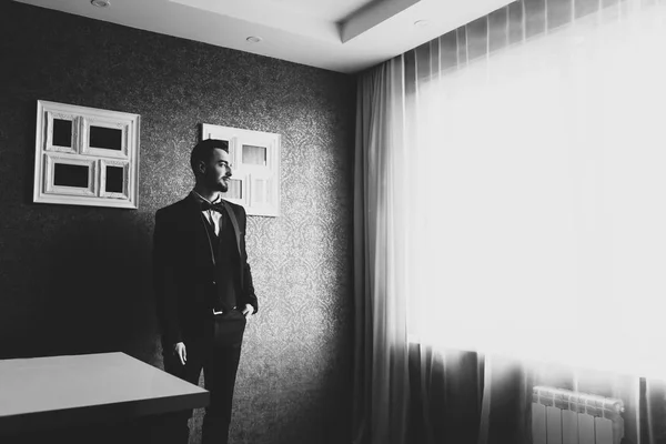 Stylish groom in white shirt and bow tie posing at window light. Confident and happy portrait of man. Groom getting ready in morning. Creative wedding photo
