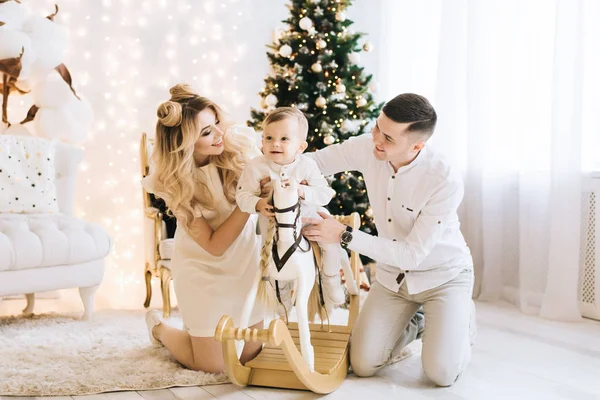 在圣诞树的背景下 一个美丽的年轻家庭的肖像 迷人的家庭庆祝新年 婴儿骑着白马和父母微笑 — 图库照片