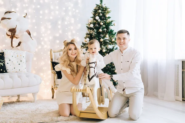 在圣诞树的背景下 一个美丽的年轻家庭的肖像 迷人的家庭庆祝新年 婴儿骑着白马和父母微笑 — 图库照片