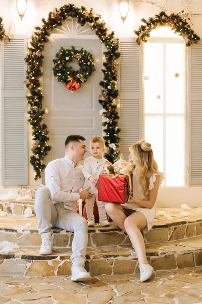 Happy young family sitting on the porch of the house, decorated for Christmas and preparing gifts for the holiday