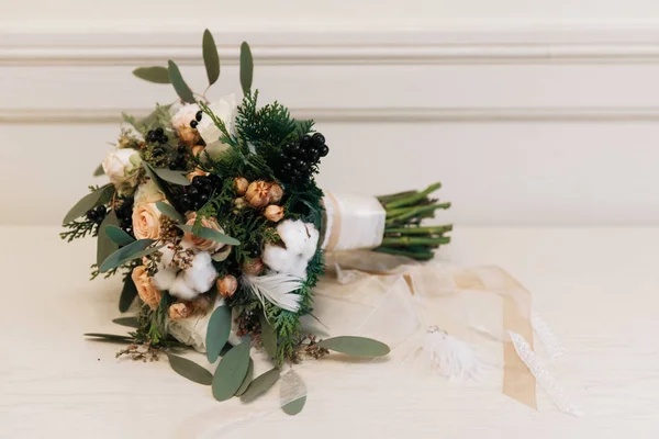 Winter bride\'s bouquet in pastel colors with roses, spruce, cotton, black berries, feathers and dried flowers. Lies alongside earrings with feathers.  Rustic. Selective focus