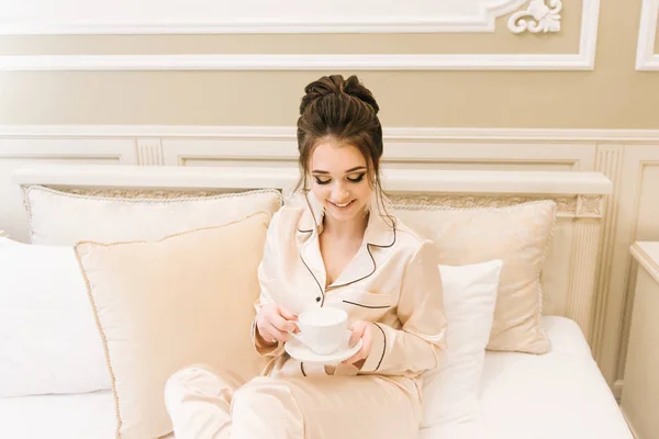 美丽的年轻女孩在金色睡衣在一个豪华的房间与时尚的头发和化妆躺在床上与一杯咖啡 早晨新娘 — 图库照片