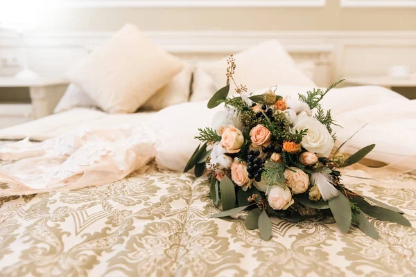 Winter bride\'s bouquet with roses, cotton, spruce lies next to the wedding dress in a luxurious hotel room