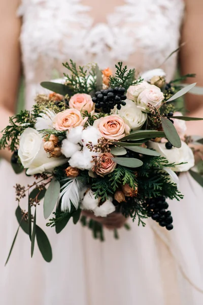 Winter bride\'s bouquet with roses, cotton, spruce, feathers, dried flowers in the hands of the bride