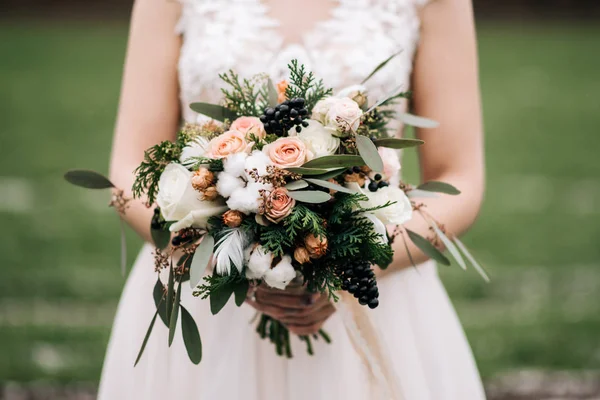Winter bride\'s bouquet with roses, cotton, spruce, feathers, dried flowers in the hands of the bride