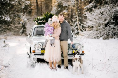 Küçük bir çocukla mutlu bir genç aile Noel için bir Noel ağacı ve hediyeler karlı kış ormandaki çatısında bir retro araba arka plan üzerinde bir husky köpek yürüyüş hazırlanıyor.