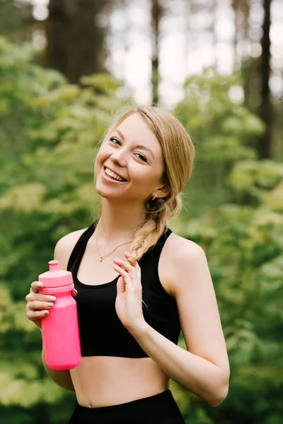 Beautiful young girl in sports uniform with a bottle of water doing yoga and sports in the forest in the open air