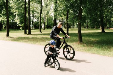 Mutlu küçük çocuk parkta genç bir babayla bisiklet sürüyor.