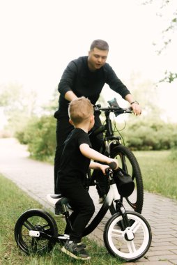 Mutlu küçük çocuk parkta genç bir babayla bisiklet sürüyor.