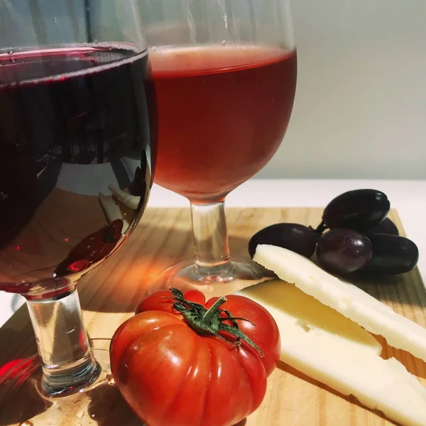 Due bicchieri di vino rosso, formaggio, uva e pomodori su una tavola di legno da cucina al sole — Foto Stock