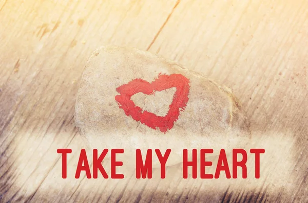 Pink heart painted with lipstick on piece of stone on background of wooden board with text Take my heart