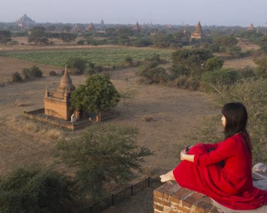 Bagan, Myanmar - 29 Ocak 2016: İsimsiz Asyalı kadın, bunlardan birinin tepesinden birçok tapınağa bakıyor. O kırmızı bir elbise giyiyor ve o taşların üzerinde oturuyor. Sabahın erken saatlerinde.