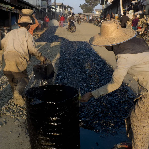 ミンデット チン州 ミャンマー ビルマ 2016 道路の砂利の上に黒いドラムからタールを広げる労働者 前景では 女性は伝統的なサロンと帽子を身に着けている — ストック写真