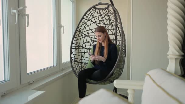 Beautiful young woman sits in a pendant chair with the phone — Stock Video