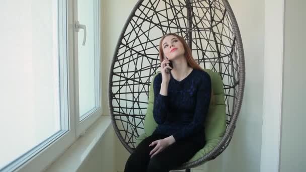 Hermosa joven se sienta en una silla y hablando por teléfono — Vídeo de stock