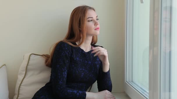 Piękna młoda kobieta wygląda przez okno, podczas gdy siedzi na parapecie w domu i marzy o czymś — Wideo stockowe