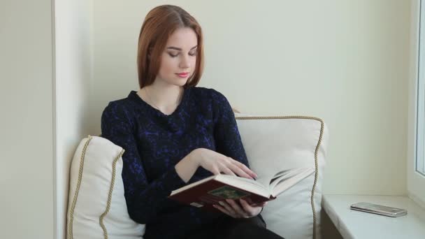 Schöne junge Frau liest ein Buch, während sie zu Hause auf der Fensterbank sitzt — Stockvideo
