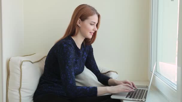 Mulher bonita se comunica em redes sociais em um laptop enquanto se senta no parapeito da janela em casa — Vídeo de Stock