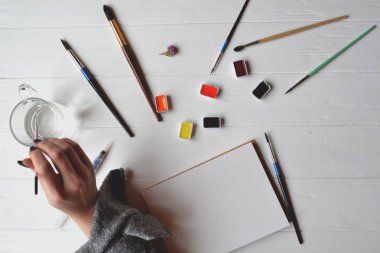 Kadın boyalar bir eskiz defteri içinde. Beyaz ahşap masa üzerinde boyama için araçlar. Ressam iş.