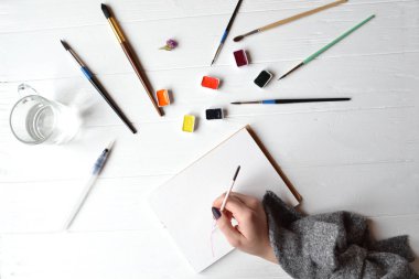 Kadın boyalar bir eskiz defteri içinde. Beyaz ahşap masa üzerinde boyama için araçlar. Ressam iş.