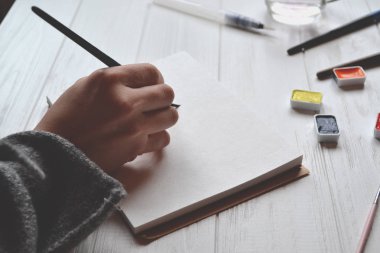 Kadın boyalar bir eskiz defteri içinde. Beyaz ahşap masa üzerinde boyama için araçlar. Ressam iş.