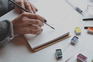 Kadın boyalar bir eskiz defteri içinde. Beyaz ahşap masa üzerinde boyama için araçlar. Ressam iş.