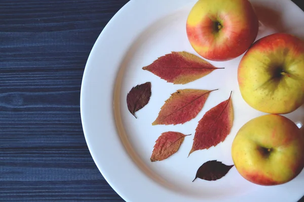 Apples in a white plate on a wooden table. Ripened apples. Autumn leaves decor.