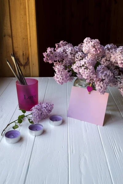 A glass with pencils, lilac in vase, empty pink paper and decorative elements on a white wooden table.