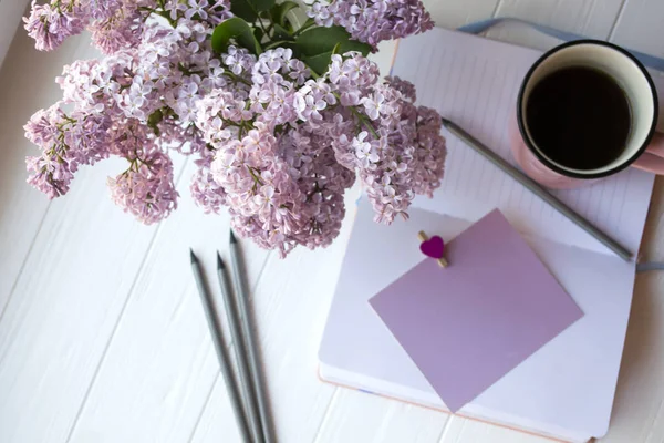 Notepad, pencils, lilac in vase and a cup of coffee on a white wooden desk. Inspirational workplace.