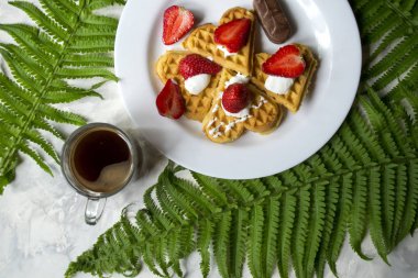 Waffle çilek ile bir fincan kahve ve yeşil fern bir masada bırakır. Güzel ve lezzetli bir kahvaltı.