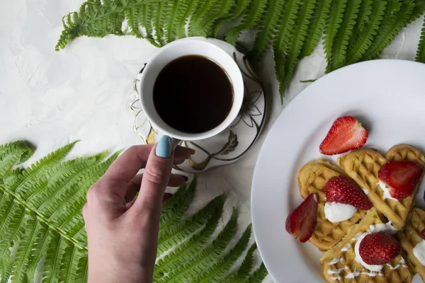 Waffles with strawberry, a cup of coffee and green leaves of fern on a table. Beautiful and tasty breakfast.