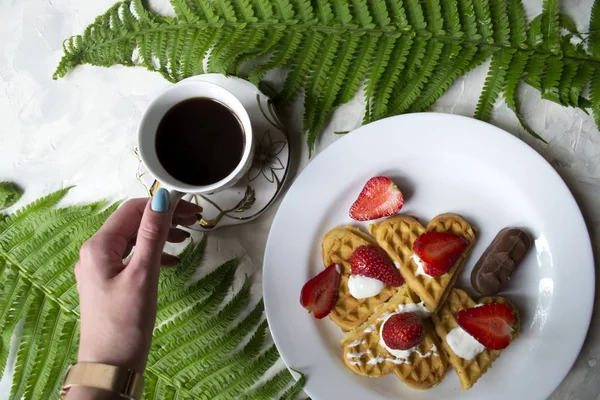 Waffles with strawberry, a cup of coffee and green leaves of fern on a table. Beautiful and tasty breakfast.