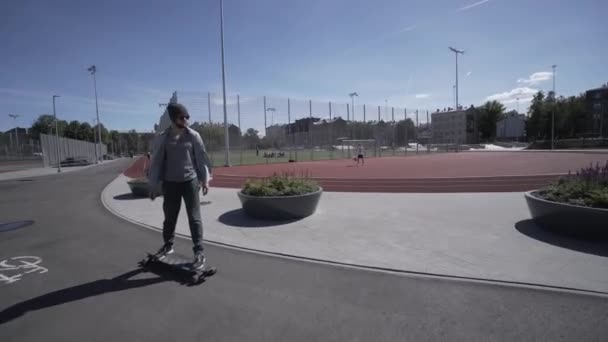 Longboard eléctrico hombre en camisa verde y paseo sombrero en el estadio deportivo rojo con patio de recreo — Vídeos de Stock