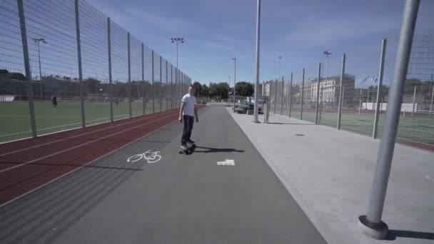 Elektrische Longboard man in wit t-shirt en een hoed rijden op rode sport stadion met speeltuin — Stockvideo