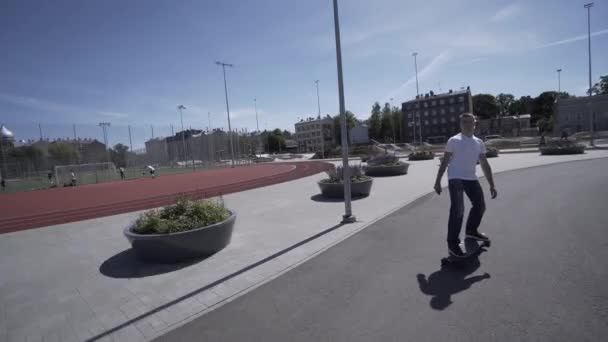 Longboard eléctrico hombre en camiseta blanca y paseo sombrero en el estadio deportivo rojo con patio de recreo — Vídeos de Stock