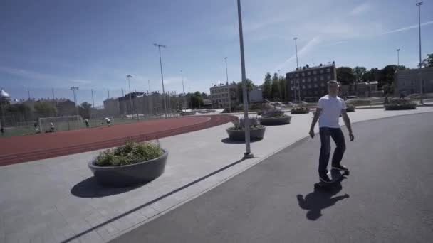 Uomo elettrico Longboard in t-shirt bianca e cappello giro sullo stadio sportivo rosso con parco giochi — Video Stock