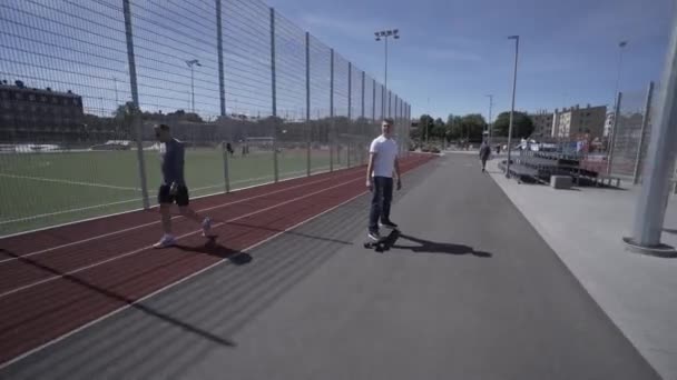 Longboard électrique homme en t-shirt blanc et chapeau tour sur le stade de sport rouge avec aire de jeux — Video