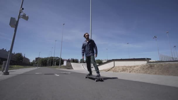 Longboard électrique homme en veste bleue et chapeau tour sur le stade de sport rouge avec aire de jeux — Video