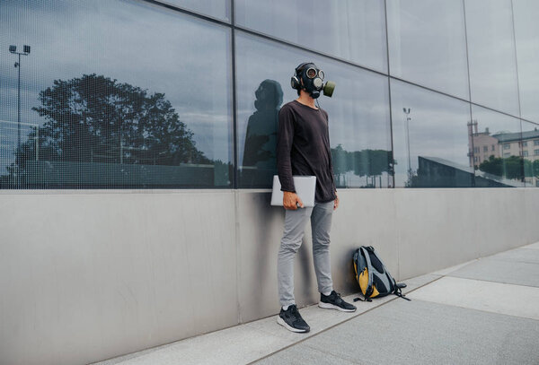 A city man in a gas mask works and listens to music against the background of a building and trees in a vacuum without people