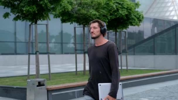 Фрилансер Мужчина, гуляющий по большому городскому городу, работает с камерой ноутбука и слушает музыку через наушники — стоковое видео