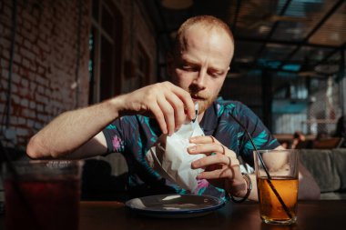 Genç adam yaz terasında hamburger yiyor.